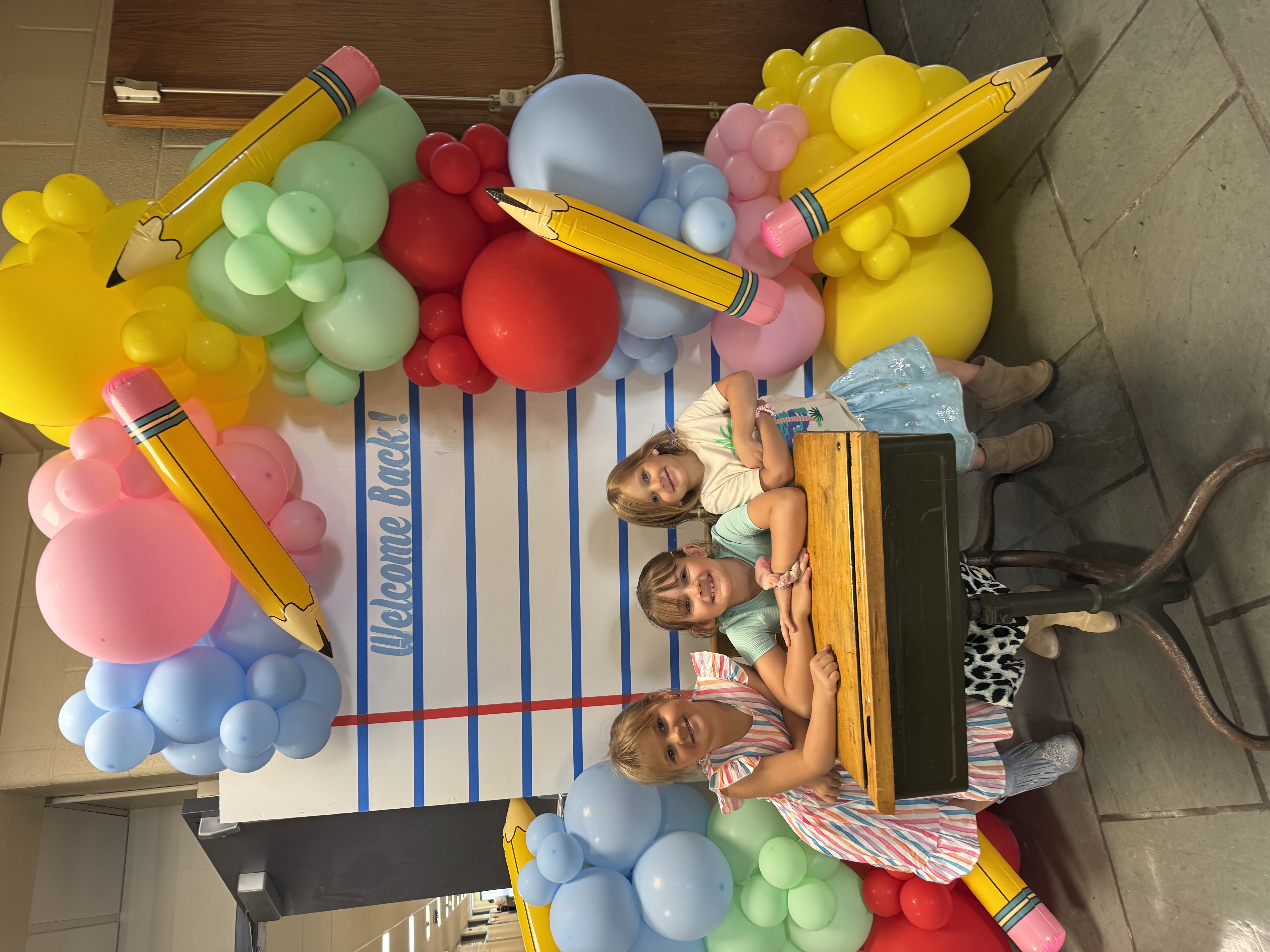 Three girls smiling in a welcome back balloon arch at MJT