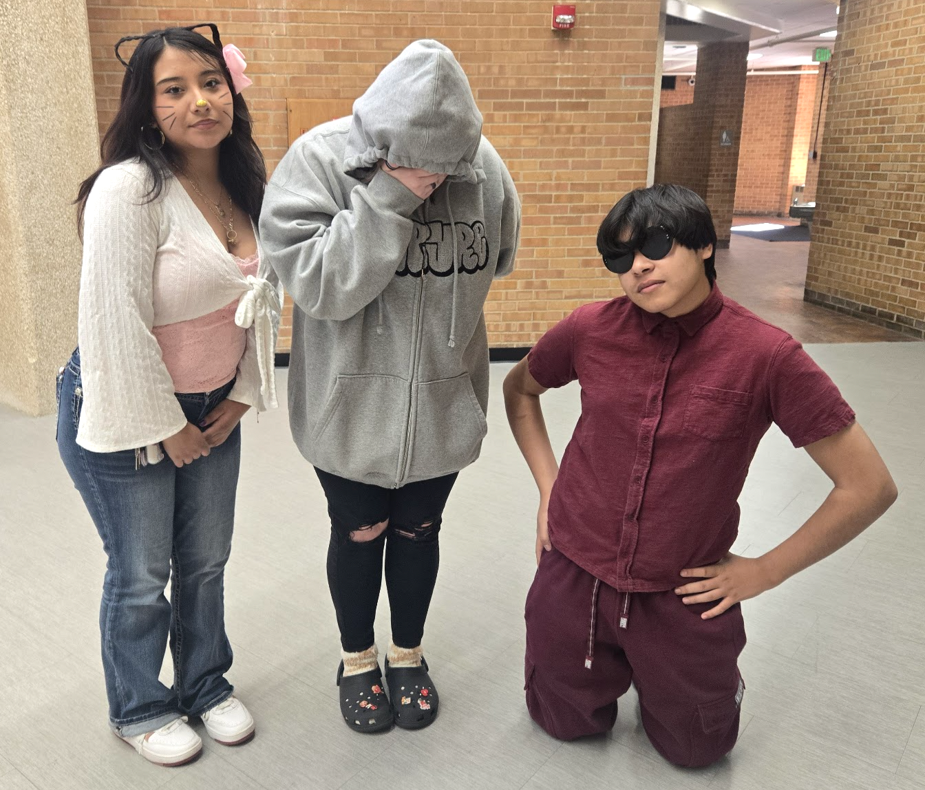 3 students standing together, one is wearing a hoodie over their face, another is kneeling on the ground with hands on hips
