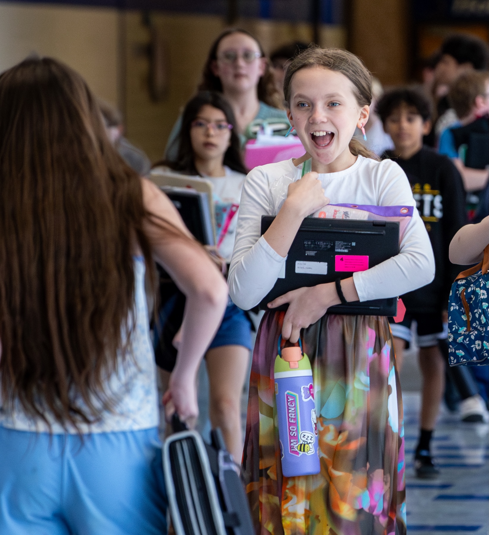 Smiling student holding a laptop and water bottle walks in a school hallway with classmates.
