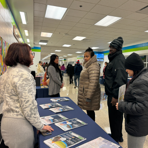 An Eastland-Fairfield staff member speaks with adults at an information table covered with Aspire program brochures during an indoor community event.