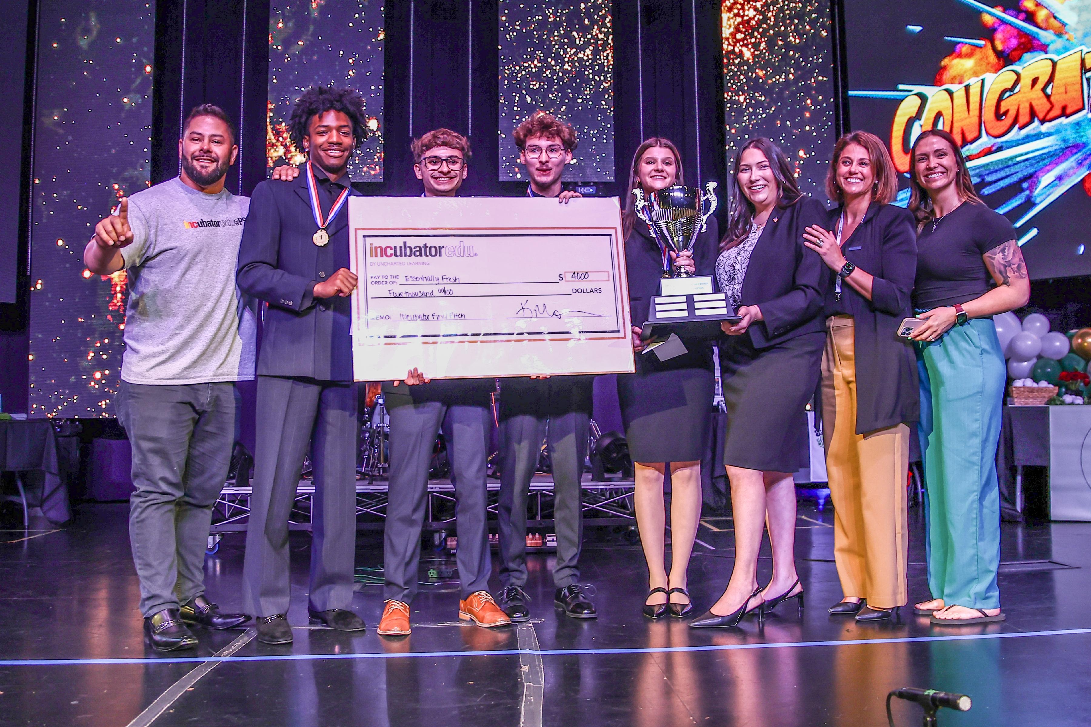 Adults and students stand on stage holding oversized check and trophy