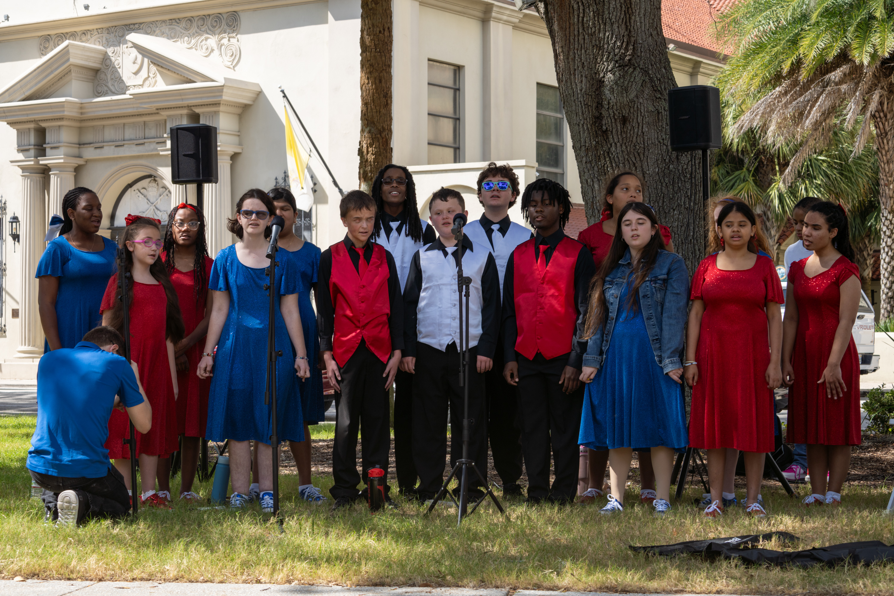Show Choir students standing in 2 lines under the shade of an oak tree. the girls are wearing red or blue glitter dresses with glitter sneakers; the boys are wearing black shirt and pants, either either a red or a white vest and tie.