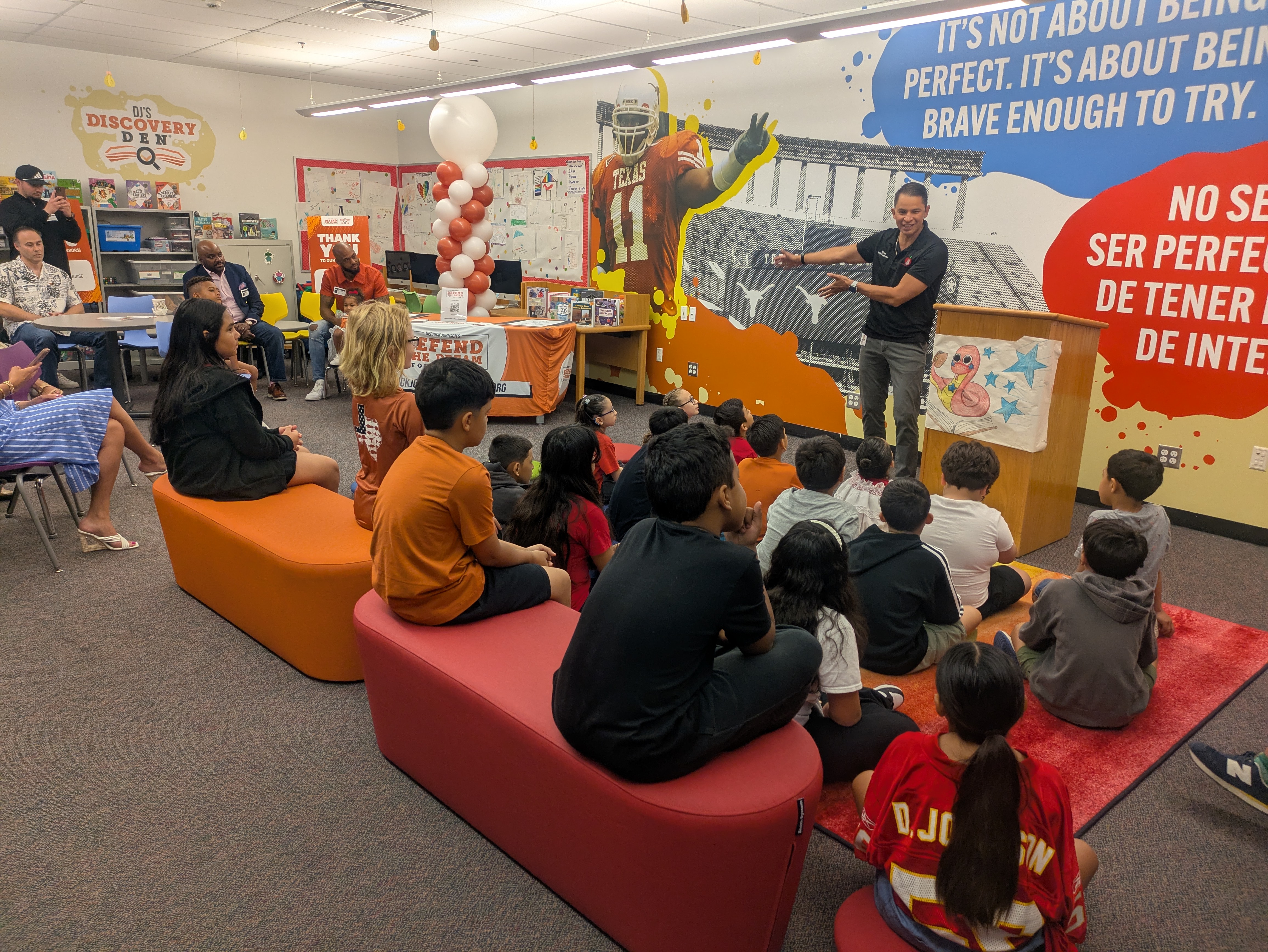 A group of elementary students sit on colorful benches and carpet, attentively listening to a speaker at a podium inside a decorated classroom. Behind the speaker is a mural featuring a football player and motivational quotes in English and Spanish. The room includes balloons, books, and signage for “DJ’s Discovery Den” and the “Defend the Dream Foundation.” Adults are seated around the room, observing the event.
