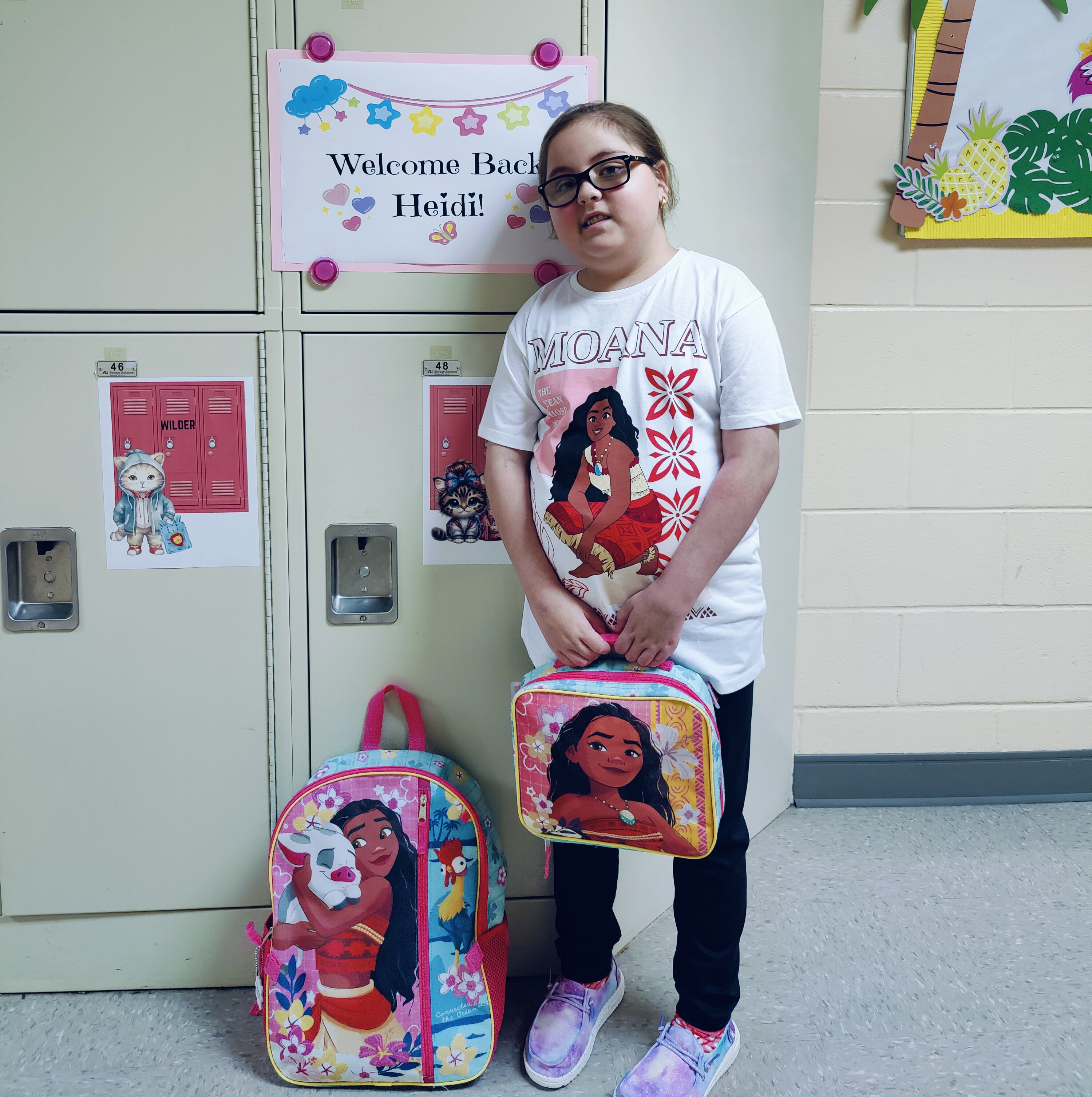 Heidi standing in front of her locker which has a poster that says Welcome Back Heidi. She is wearing a Moana-themed shirt, backpack, and lunchbag