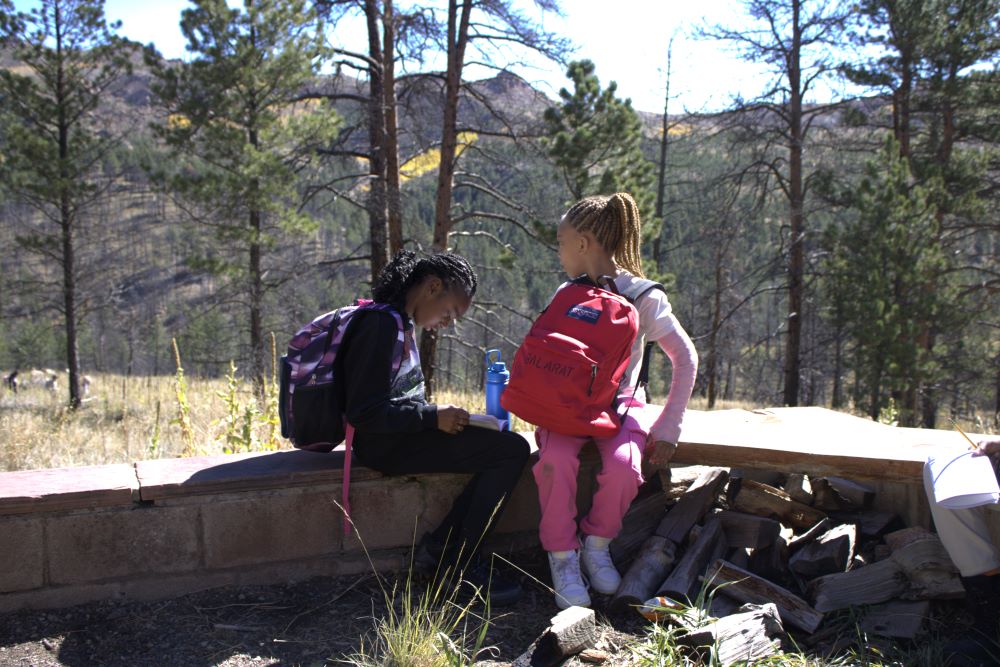 Hallett students sit outside on a bench with a view of mountains and forest behind them. One student writes in a notebook while another looks at the view.