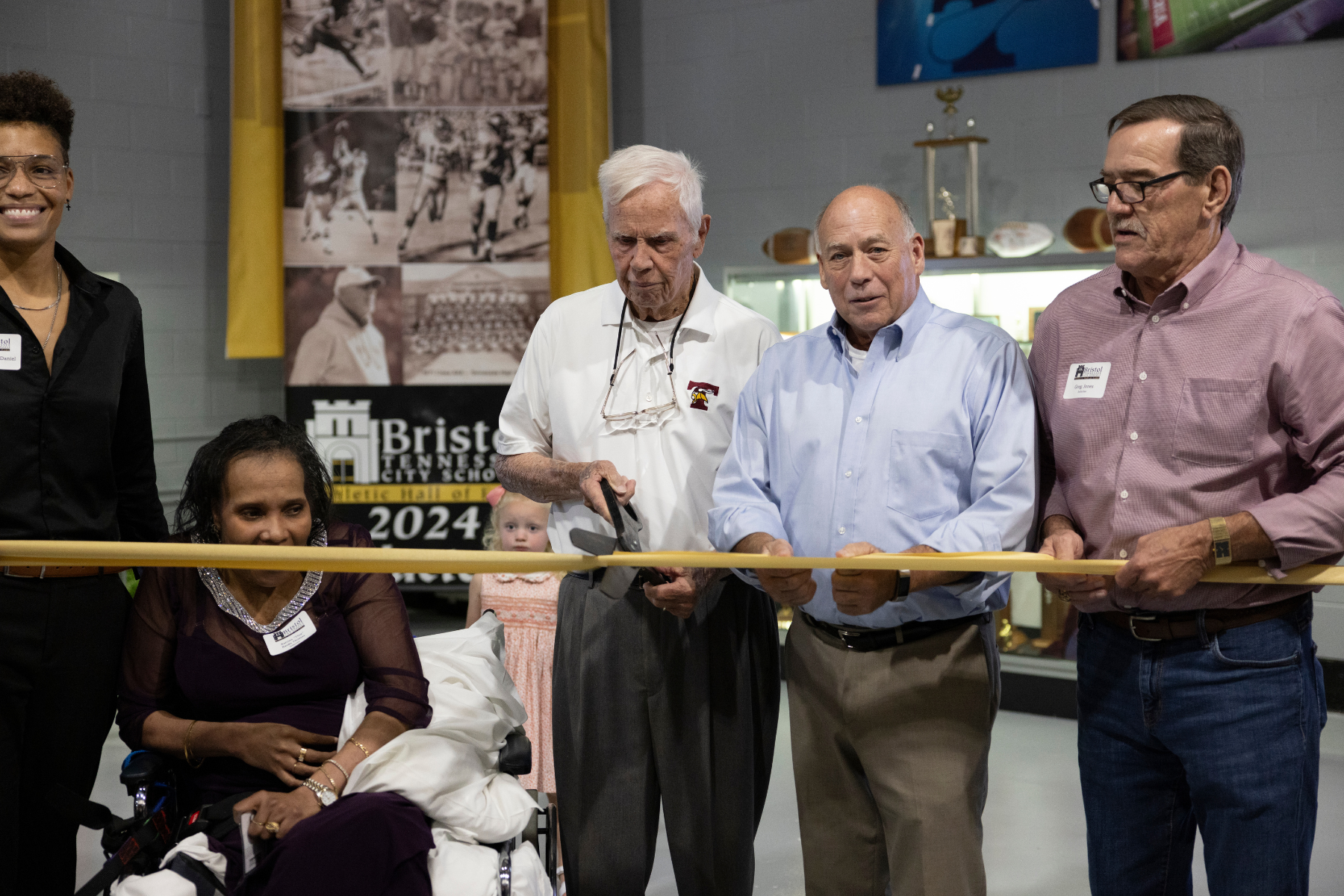 Photo of two women and three men cutting a gold ribbon in front of the Bristol Tennessee City Schools Athletic Hall of Fame. There is a small child in the background.