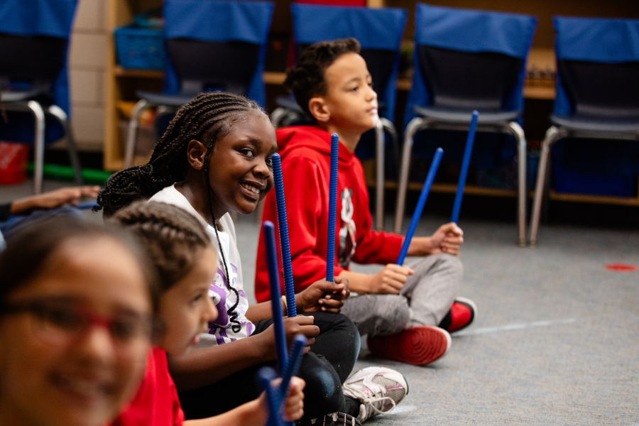Students sitting in a row on the floor as they play with musical sticks.