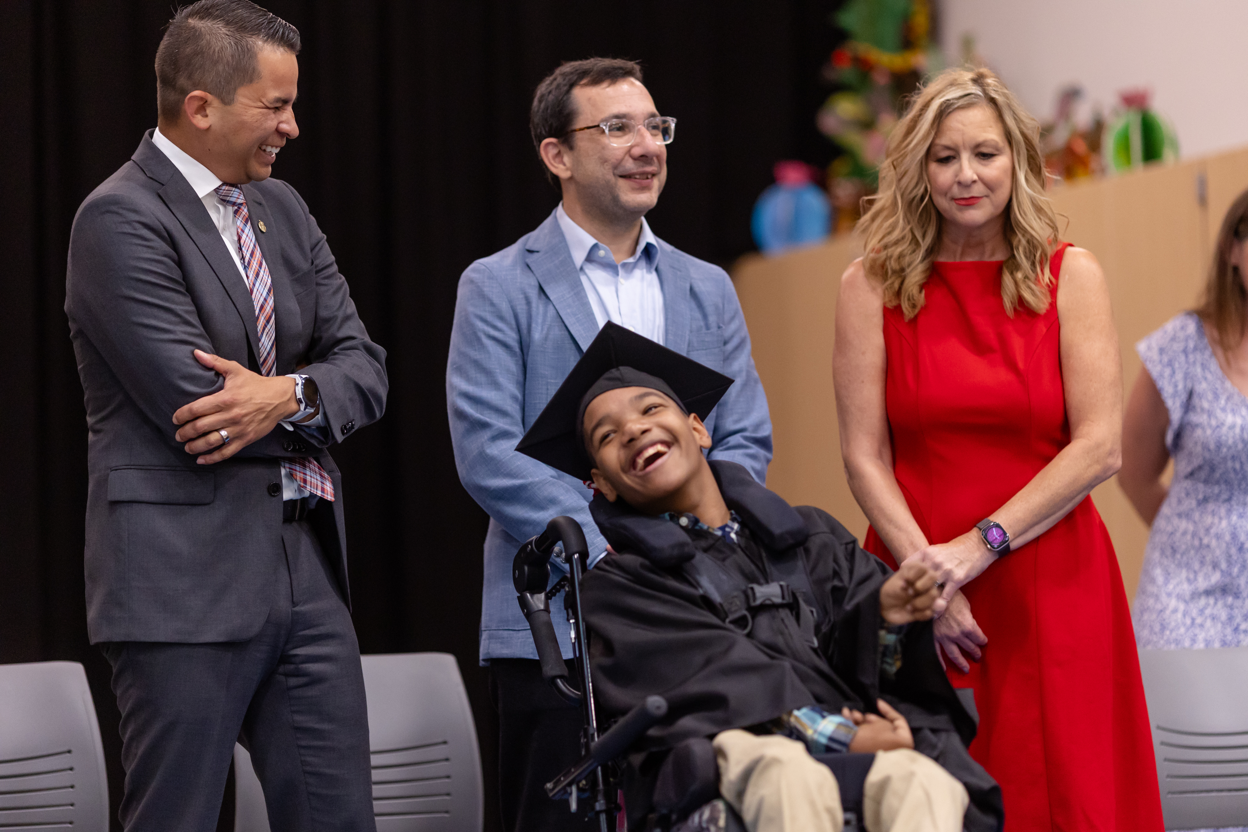 A smiling student in a graduation cap and gown sits in a wheelchair, surrounded by three adults who are also smiling and celebrating. One man in a suit is laughing with arms crossed, while another man and a woman in a red dress stand beside the student, sharing the joyful moment. The background features a dark curtain and colorful decorations.