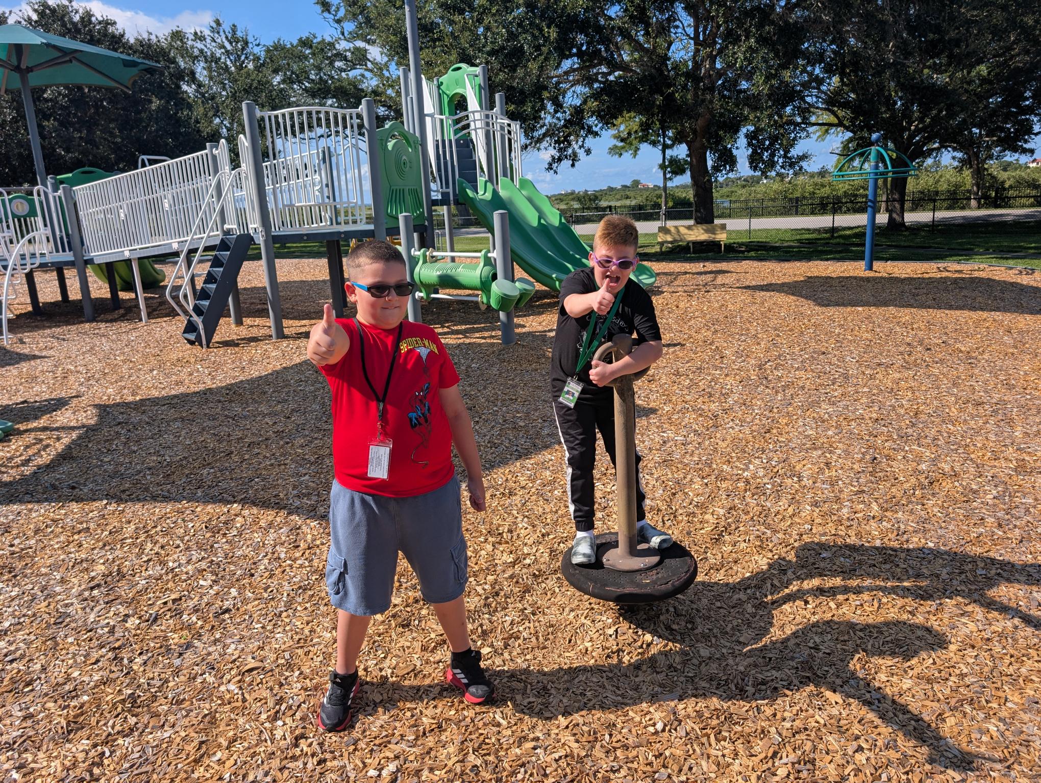 Lucas and Wilder in the mulch on playground. They are both smiling and holding thumbs up