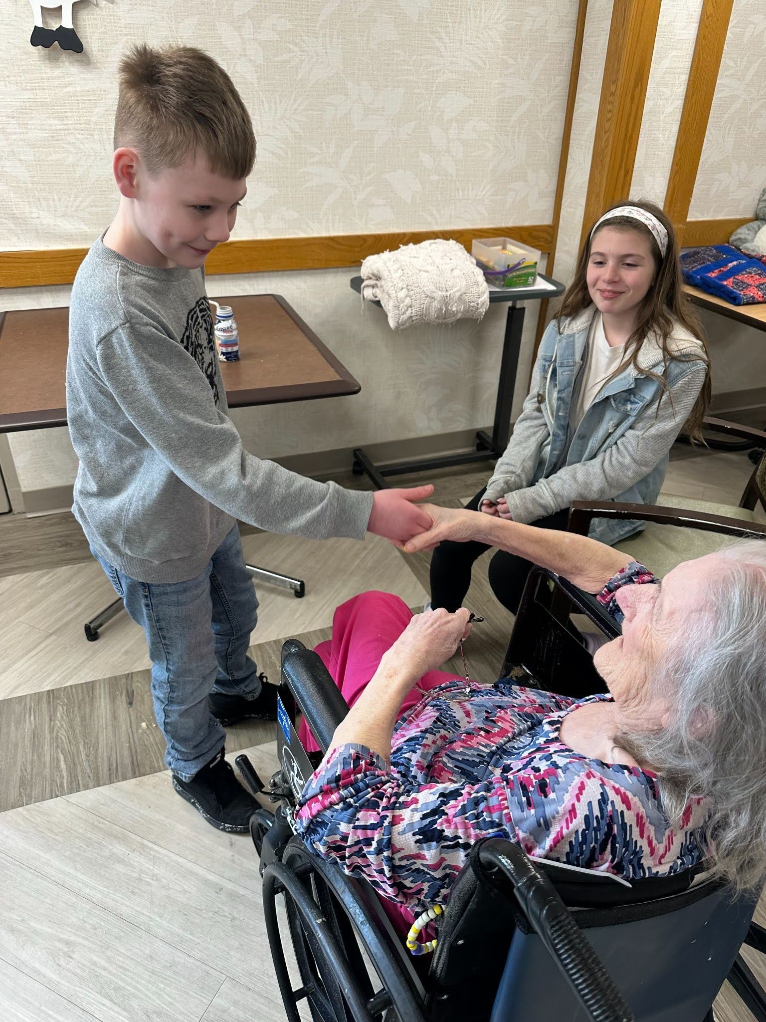 A young student is shaking the hand of an older woman in a wheel chair while a second student looks on.