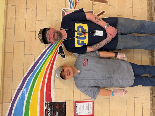 Two employees stand in front of a rainbow mural smiling.