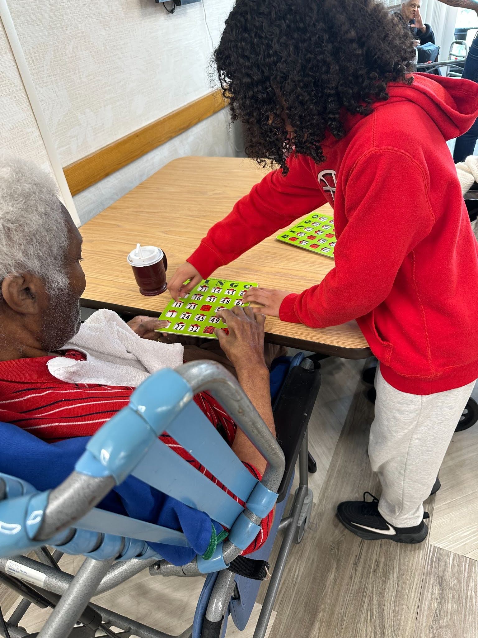 A student in a red sweatshirt stands next to an older seated man helping him to play bingo.