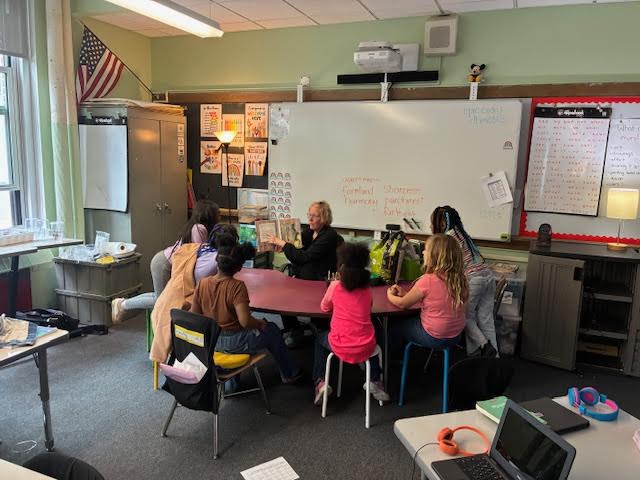Students sit a table looking at a teacher holding up a book.