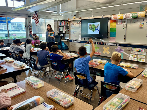Teacher leads a classroom lesson as students raise hands to participate.