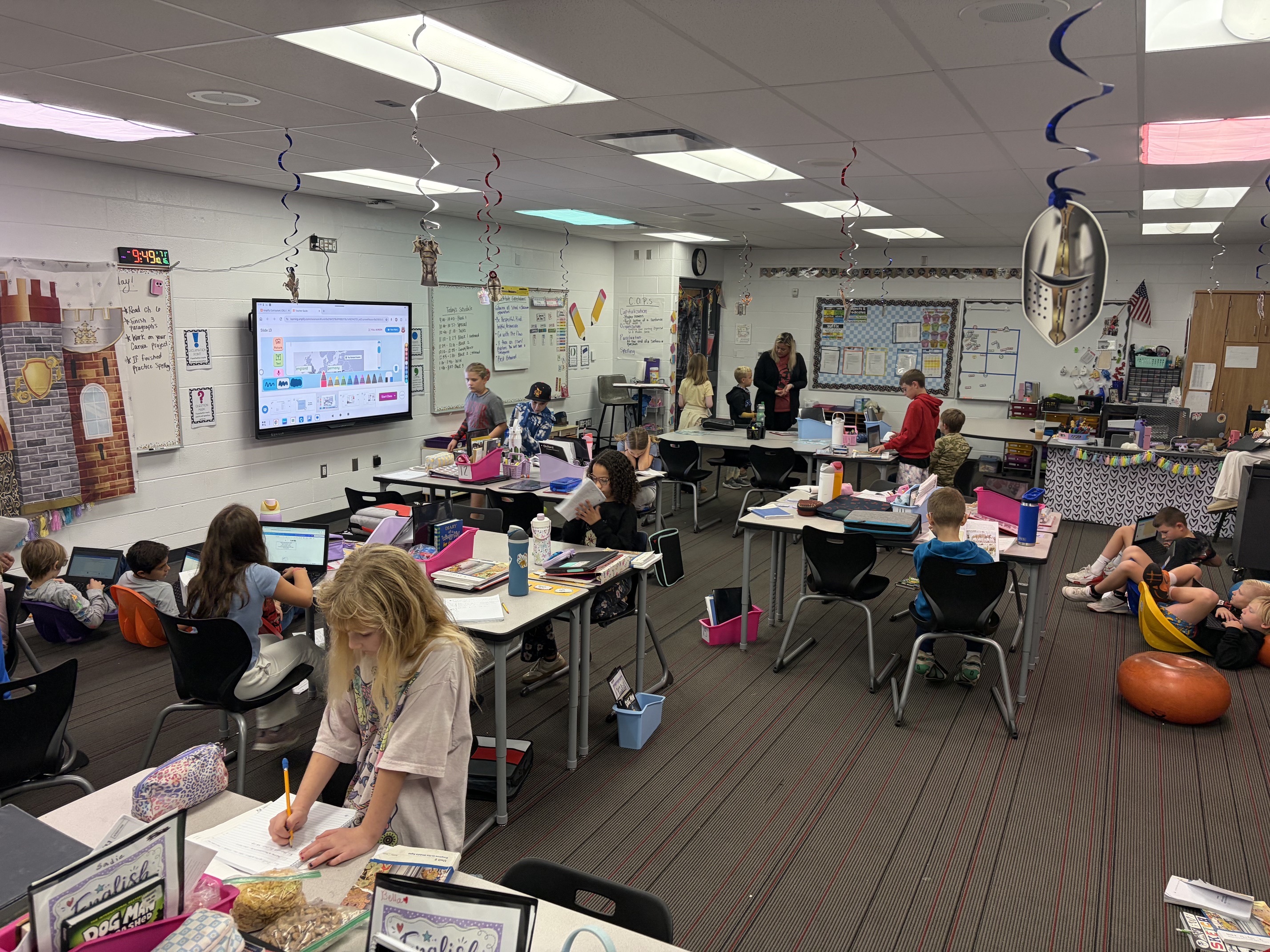 A classroom with tables and chairs, children sitting, working on computers, and a teacher at a front screen.