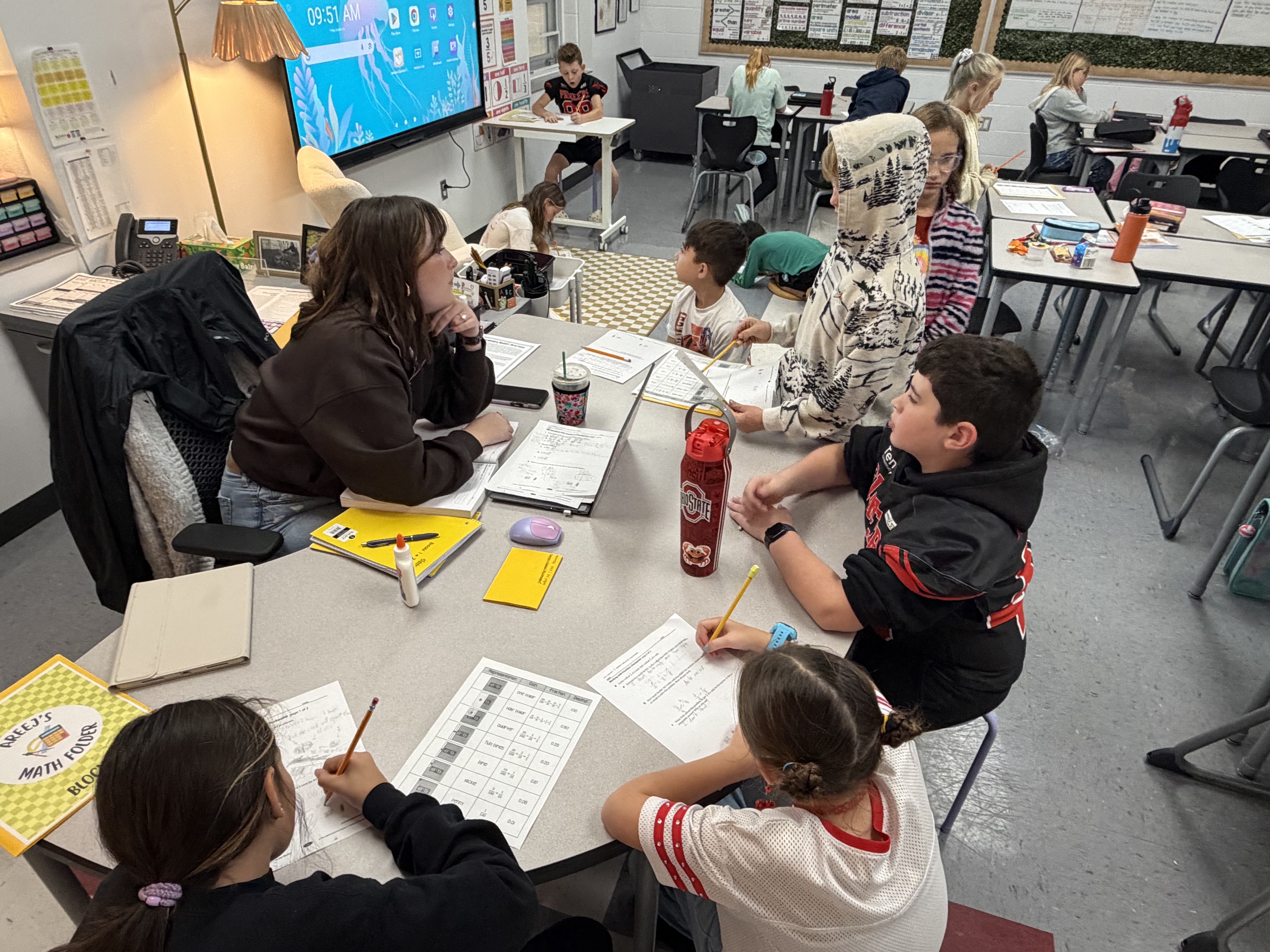 Students sit around a table with writing materials; some use pencils, others hold cups. Behind them, a classroom has tables and chairs.