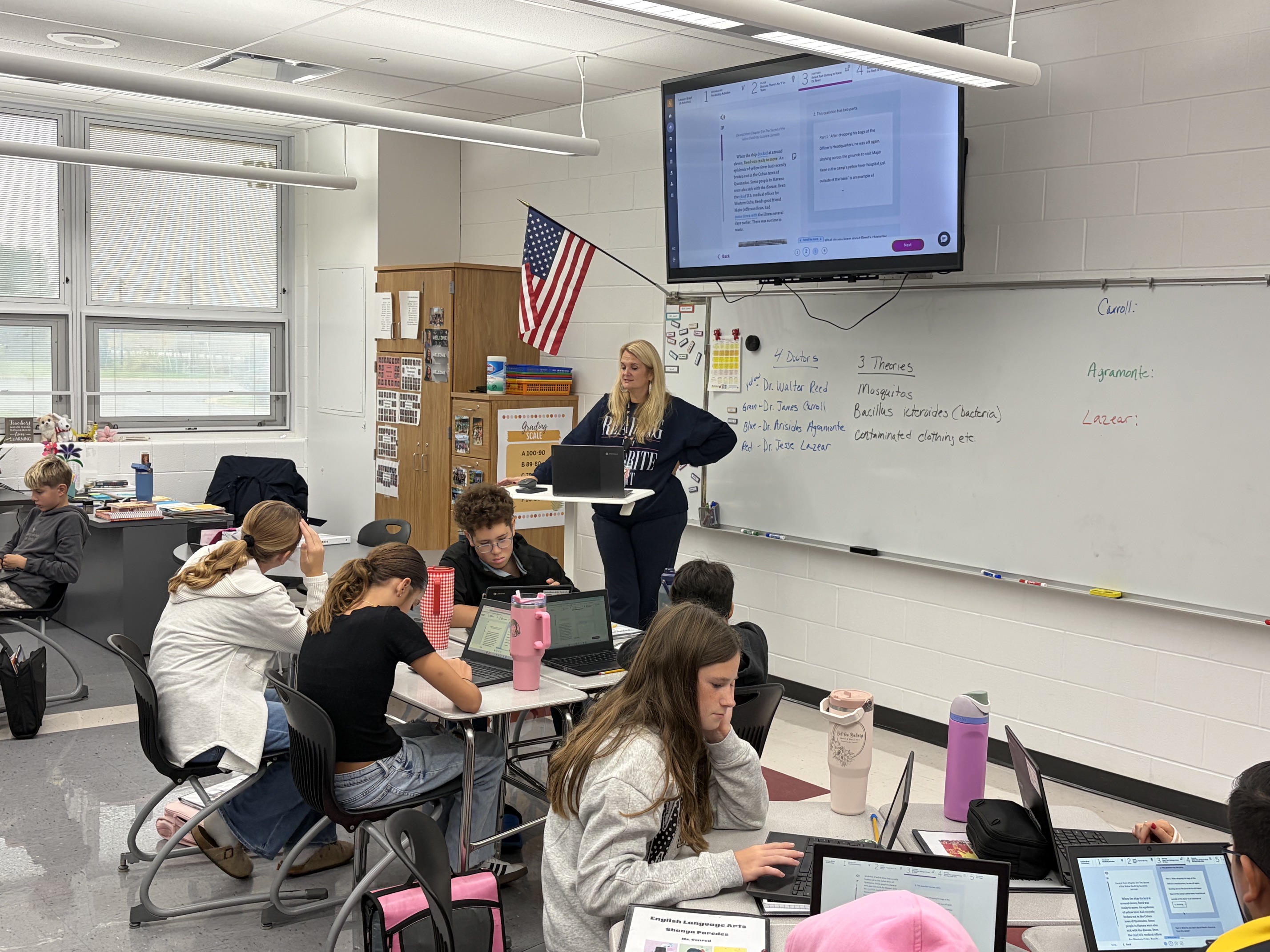 Classroom with a teacher standing by a whiteboard. Students seated at tables using laptops and other devices.