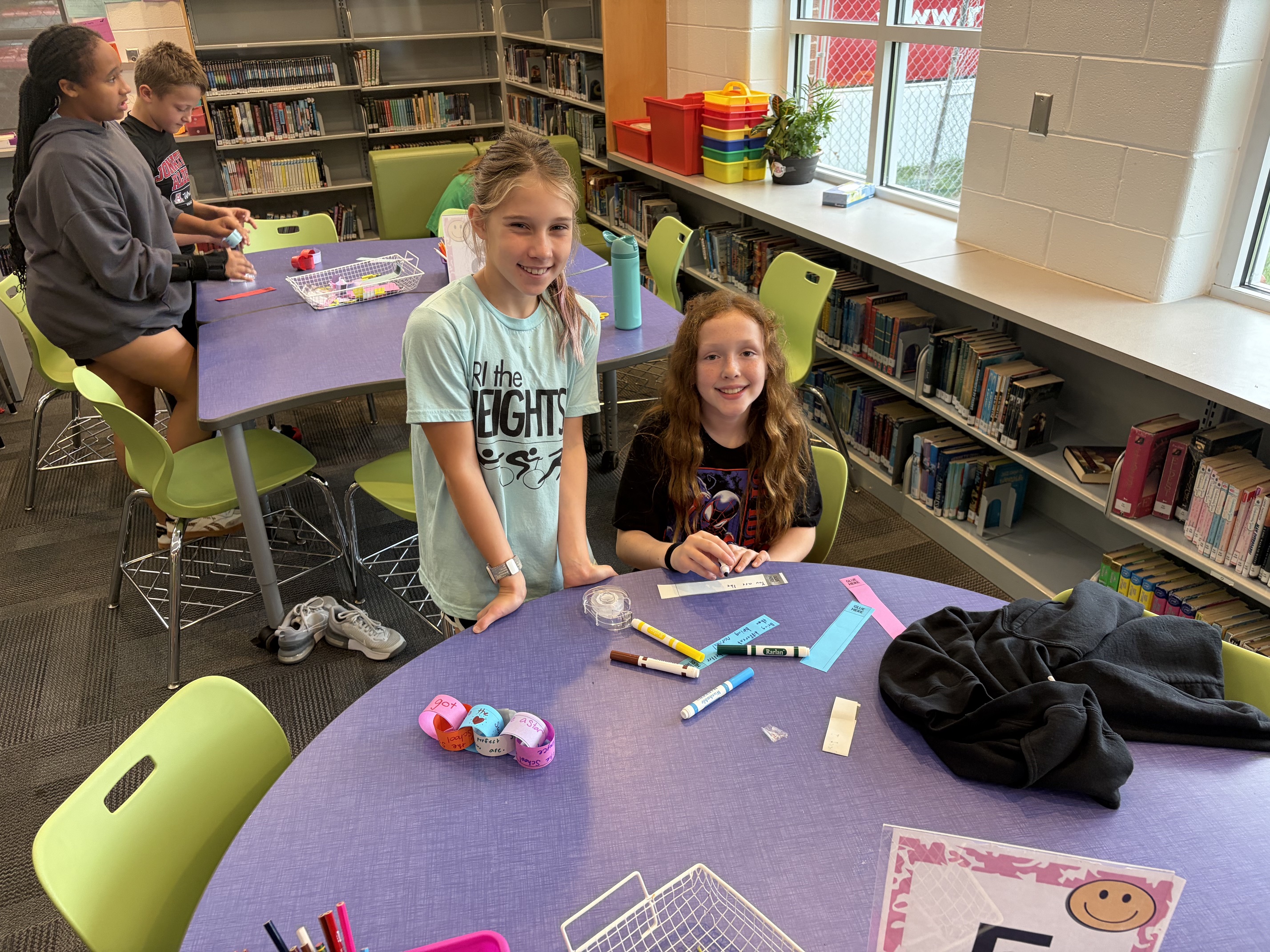 Two girls in a library. One is holding markers and the other has a jacket on a table.