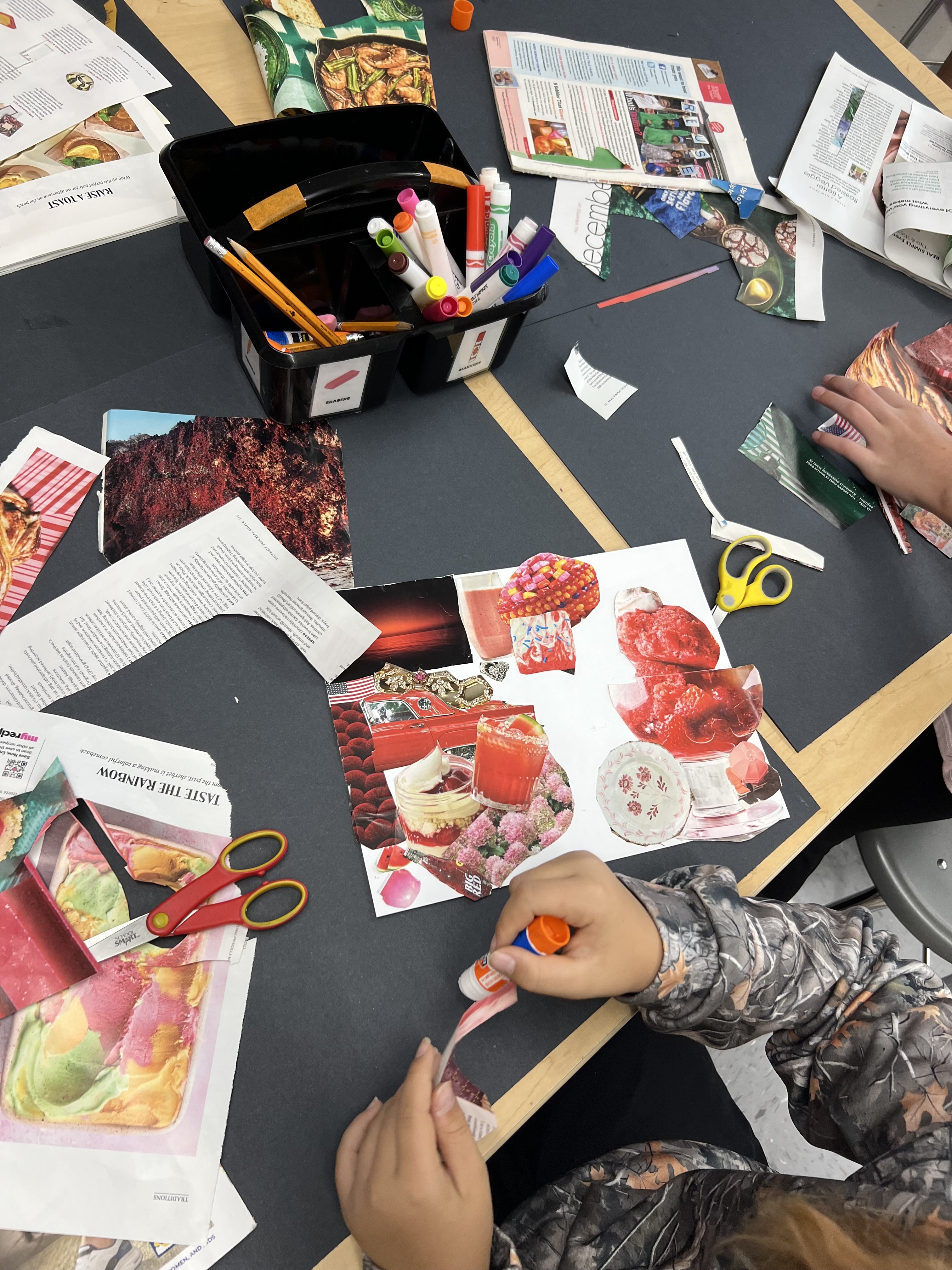 Table with various cut papers, scissors, markers, and glue. Students are working on an art project.