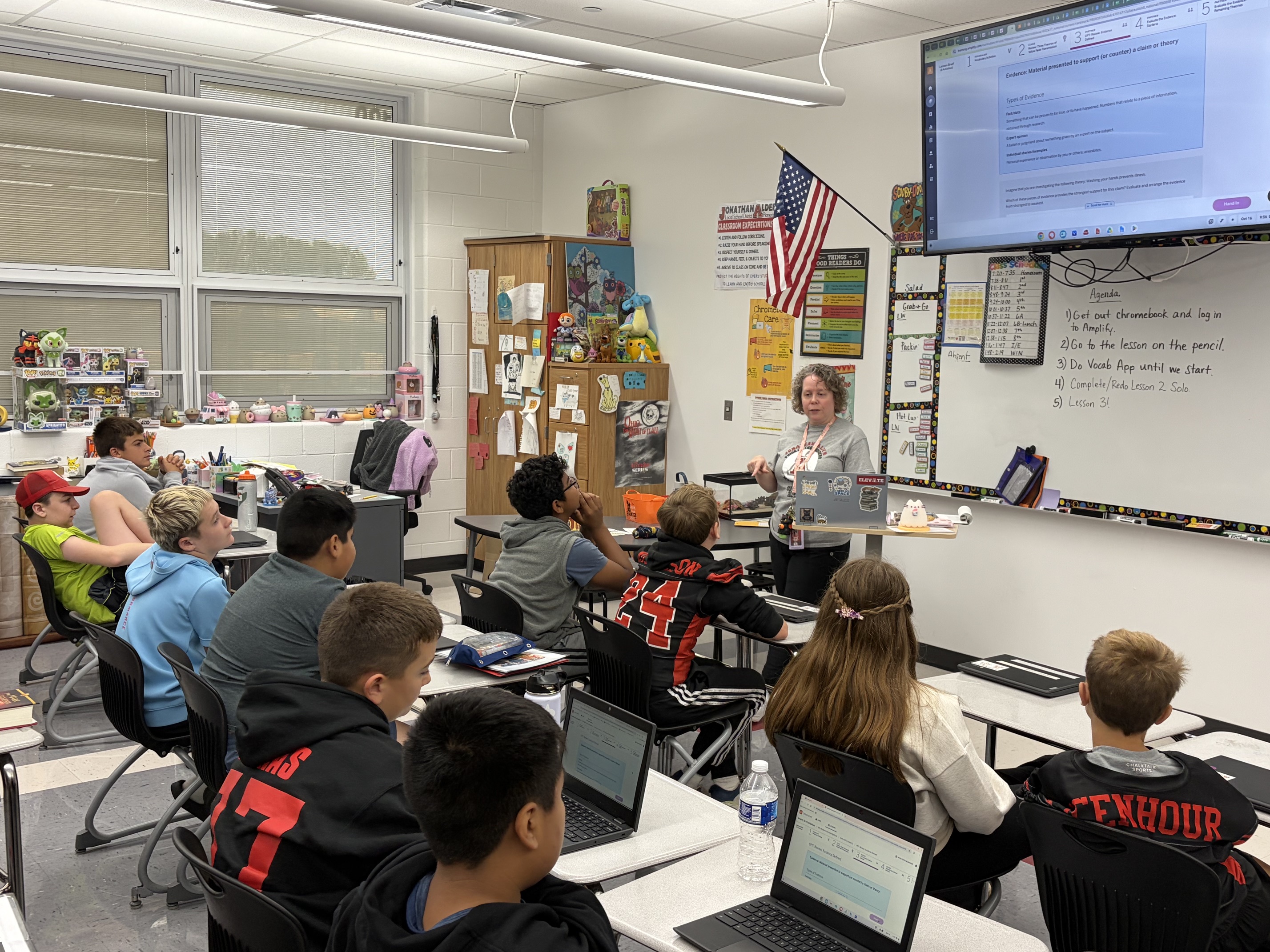 Students seated in rows of chairs around tables, with laptops, listening to a woman standing by a whiteboard.