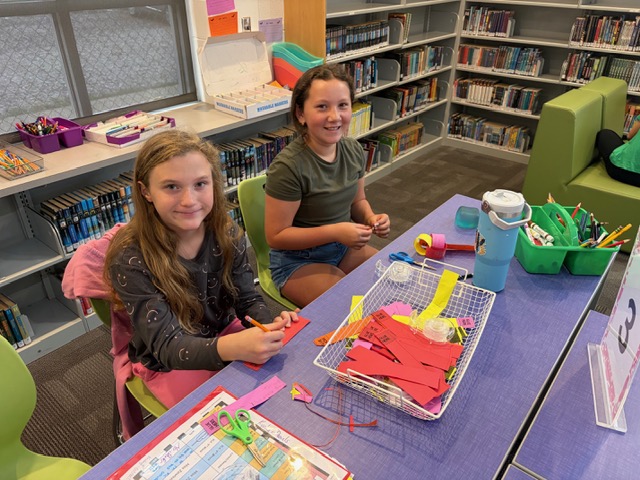 Two girls sitting at a table with art supplies, one holding a red paper, in a room with shelves.