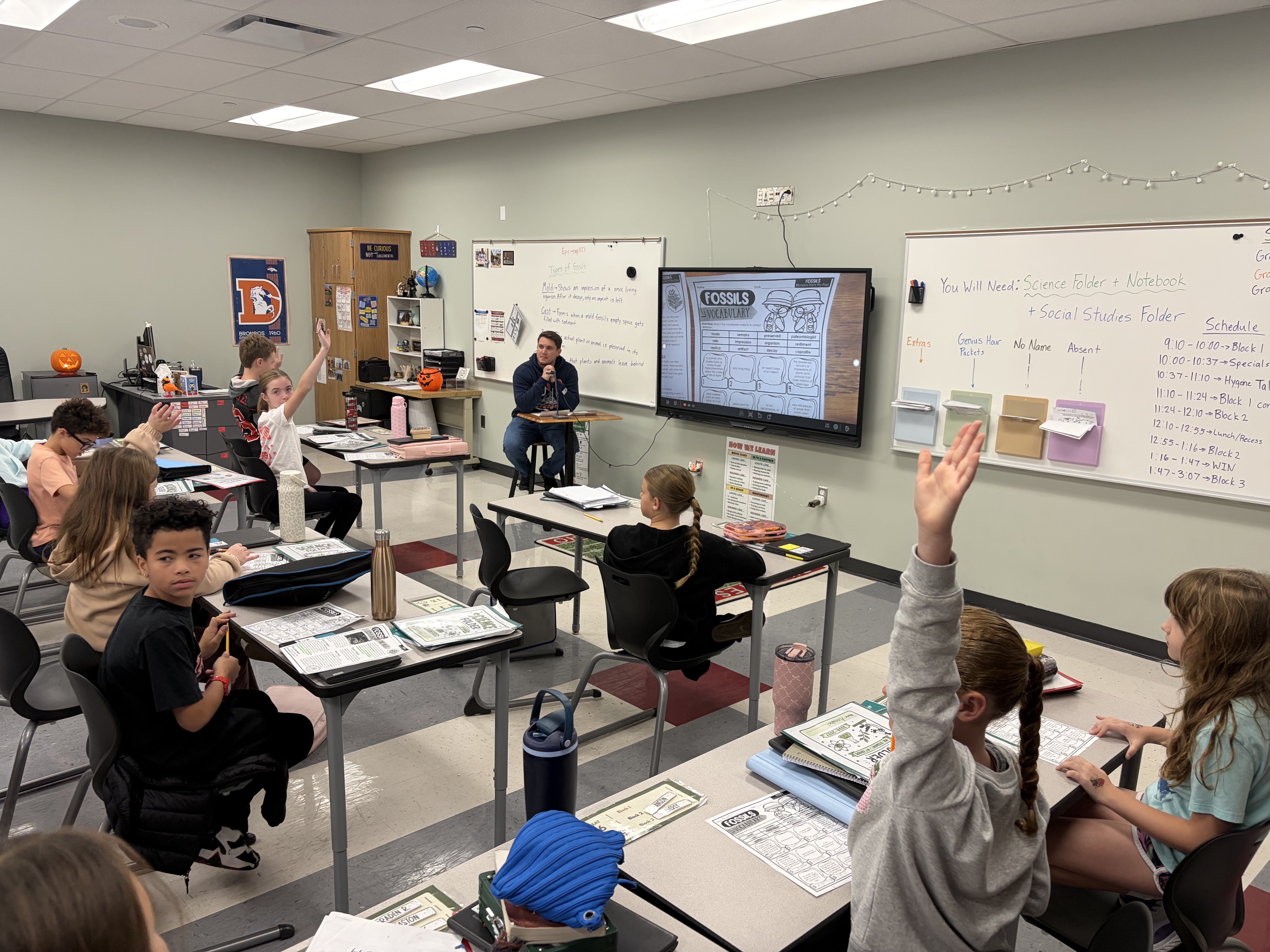 Classroom with rows of desks; students seated, some raising hands. A teacher in front of a screen.