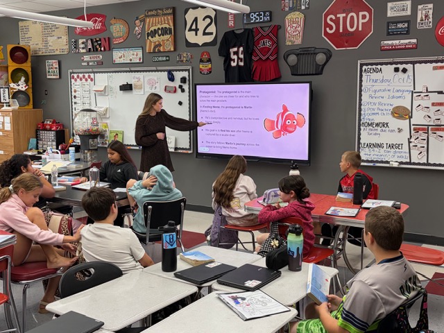 Classroom with children sitting at desks. A teacher stands in front of a projector screen showing a diagram.