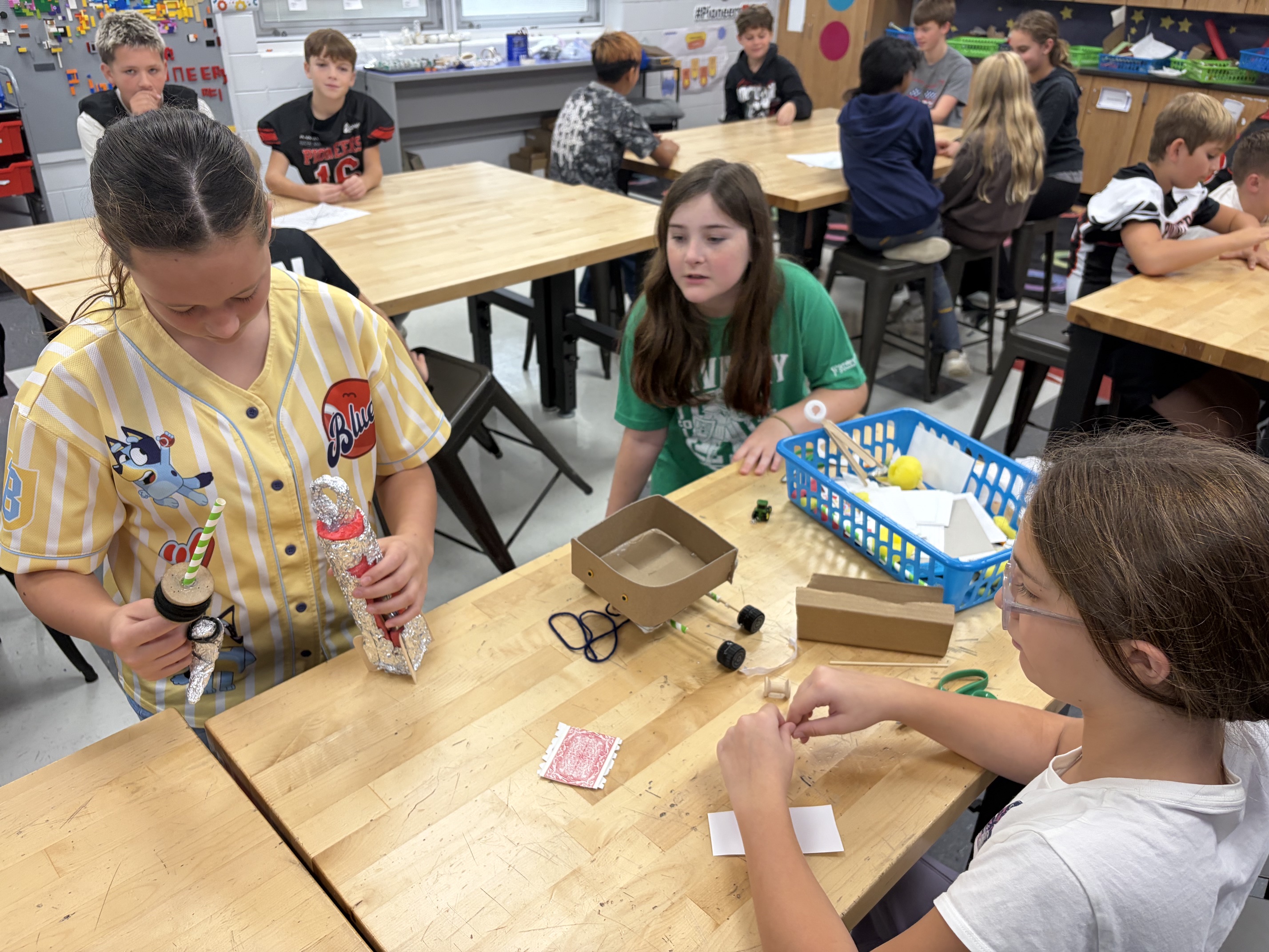Students seated at tables in a classroom, one holding a bottle, another cutting with scissors.