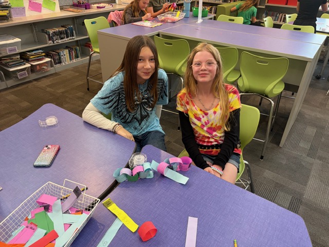 Two girls in a library, sitting at a purple table, with paper and paper craft tools.