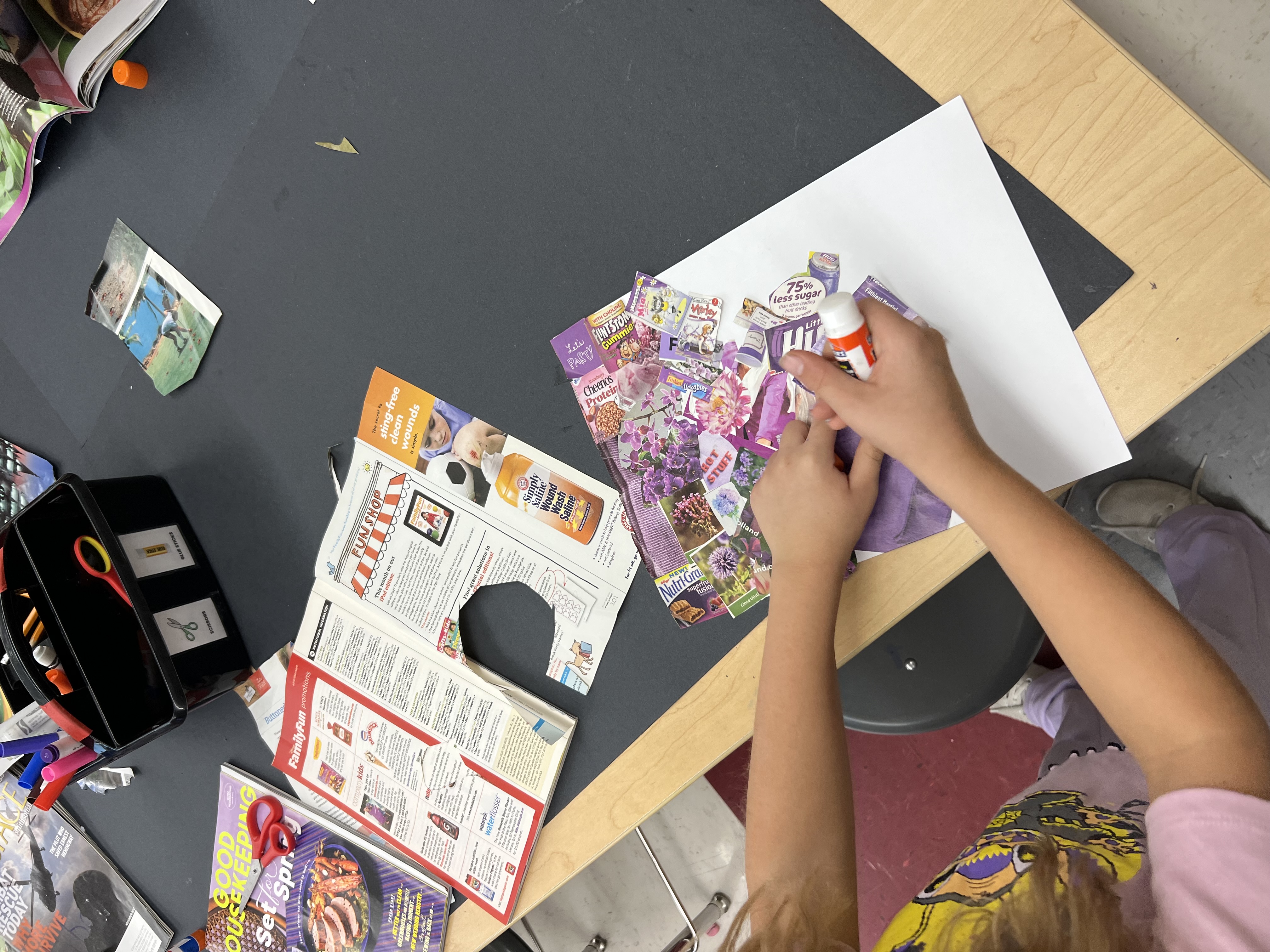 Person sitting at a table, working on a collage with glue, paper, and various printed materials.
