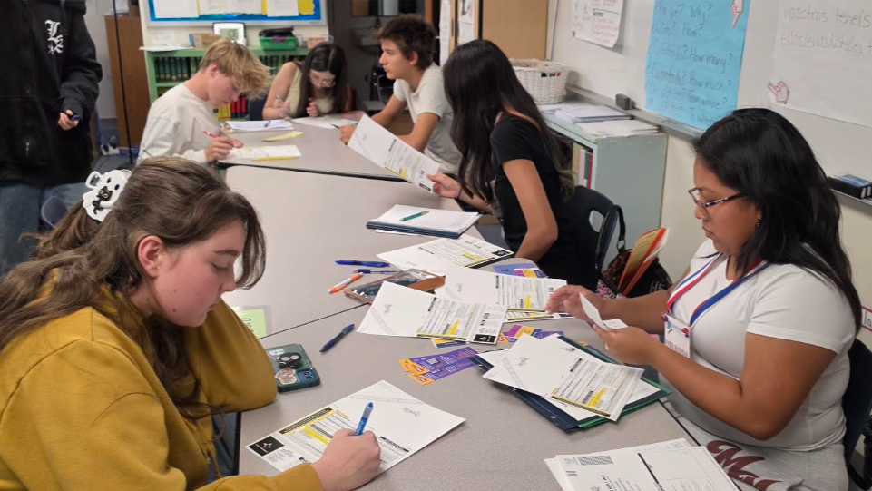 Students work on papers and writing at a long table in a classroom with whiteboards in the background.