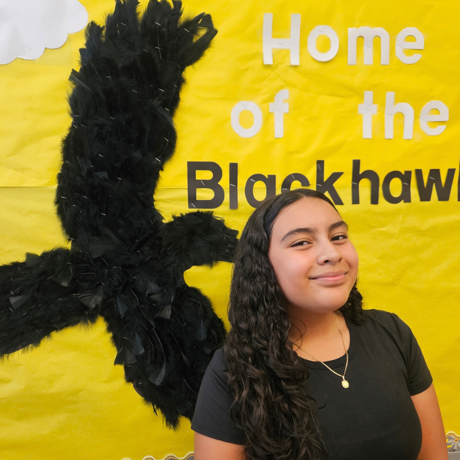 A girl with curly hair smiles in front of a backdrop with a large black feather and text.