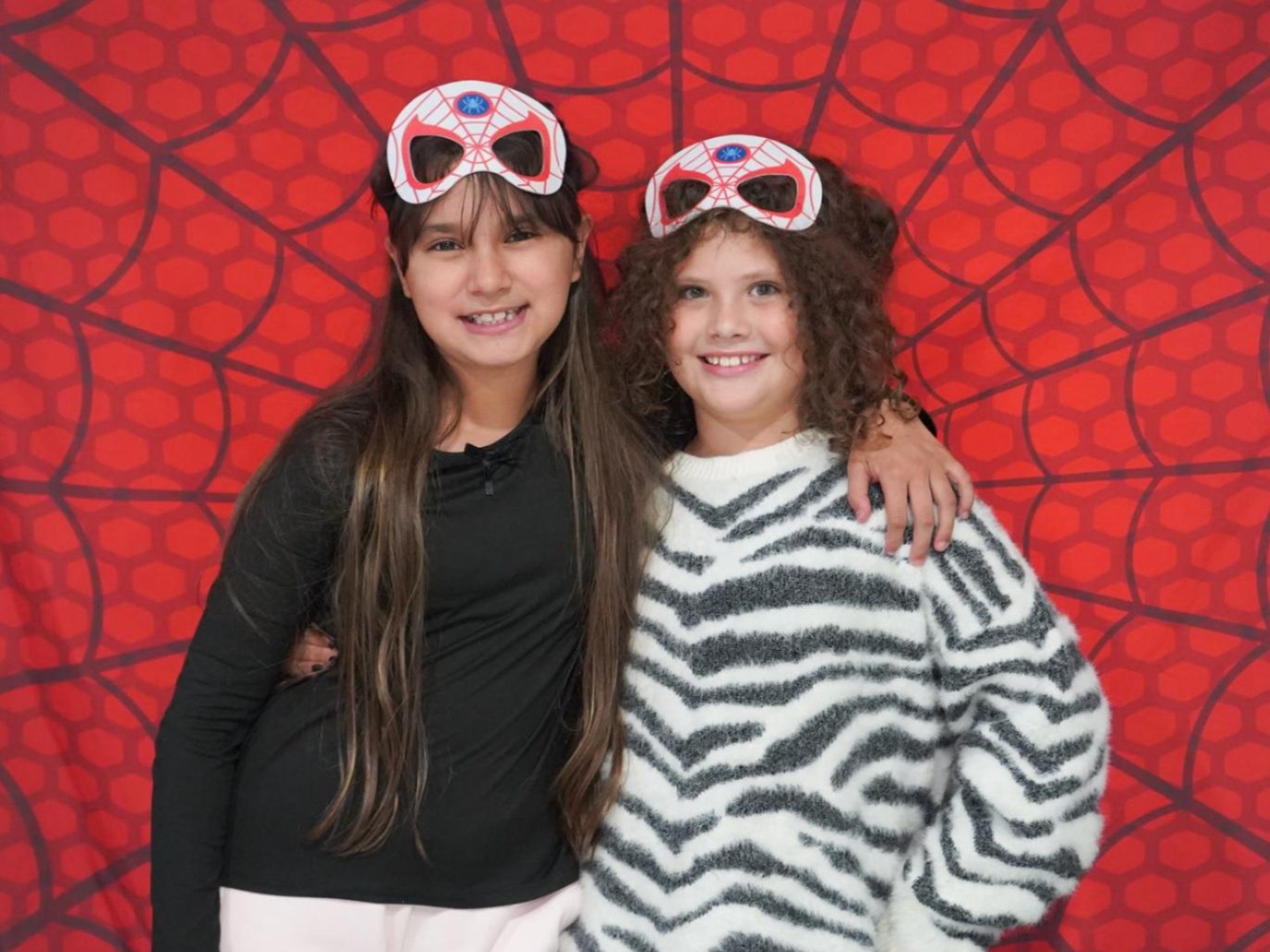 Two girls smile and pose in front of a red background with a spider-like pattern.