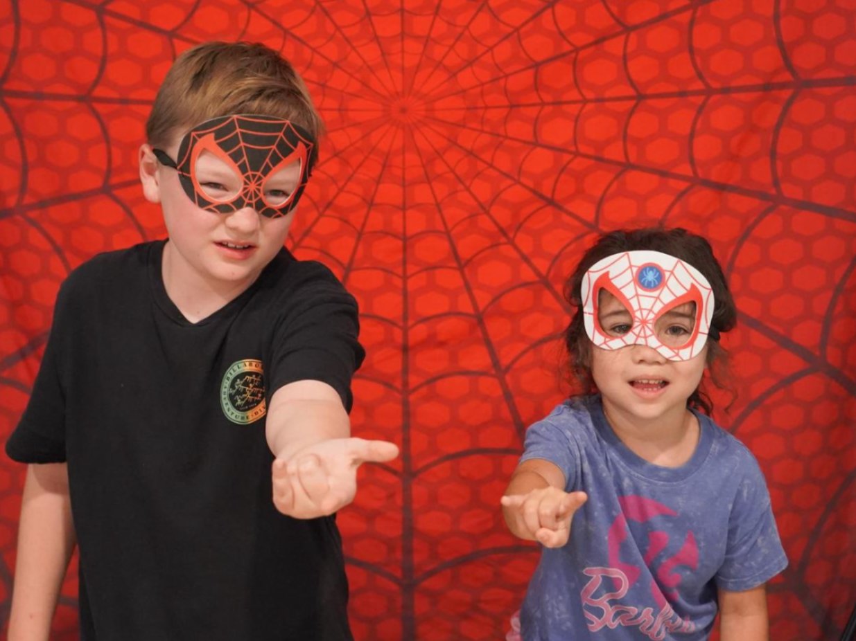 Two children stand against a red backdrop with Spider-Man designs. Both wear themed masks, one with a spider pattern.