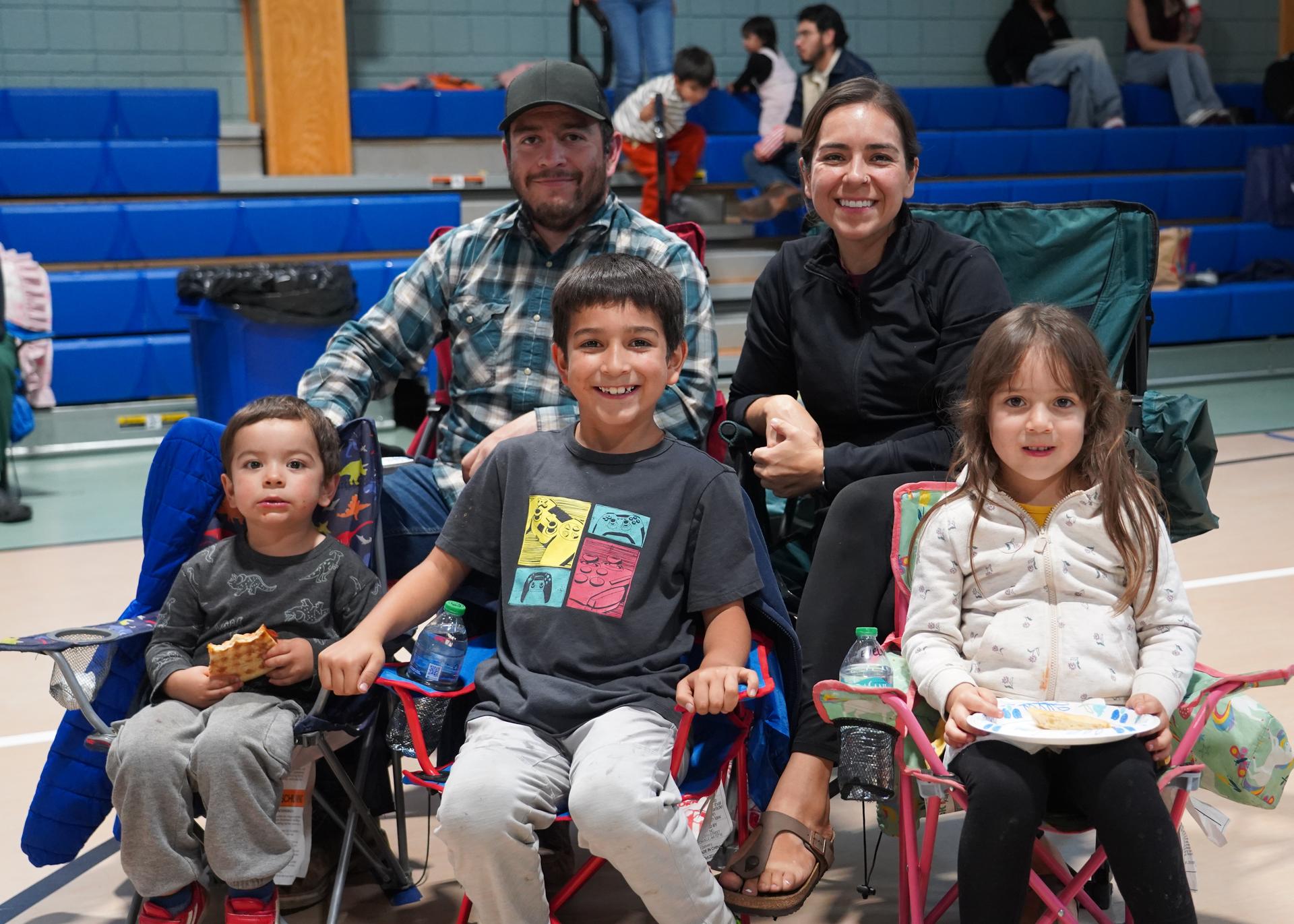 A family of four sitting in chairs in an indoor sports arena. A woman and three children.