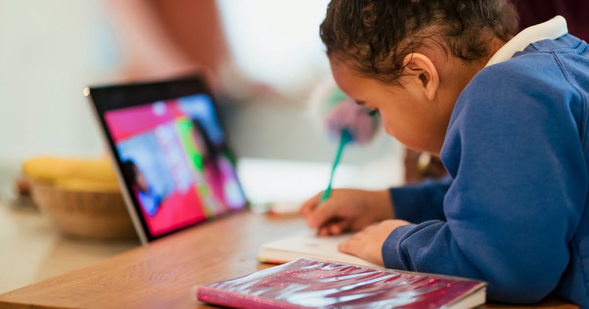 Child sitting at desk, focused on a laptop screen, holding a pen and writing in a book.