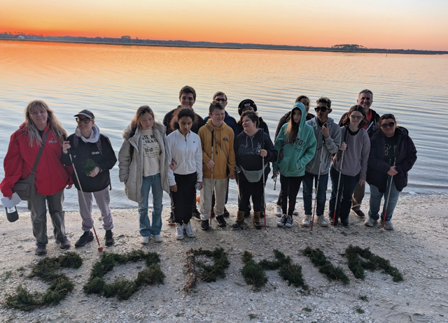 students pose with SOPHIA spelled out in seaweed on the sand in front of them, with a sunset sky and water behind them
