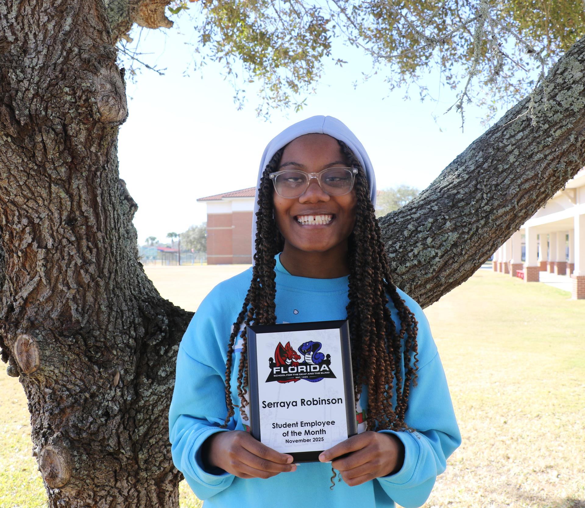 Serraya standing outside under a tree, smiling and holding her award plaque