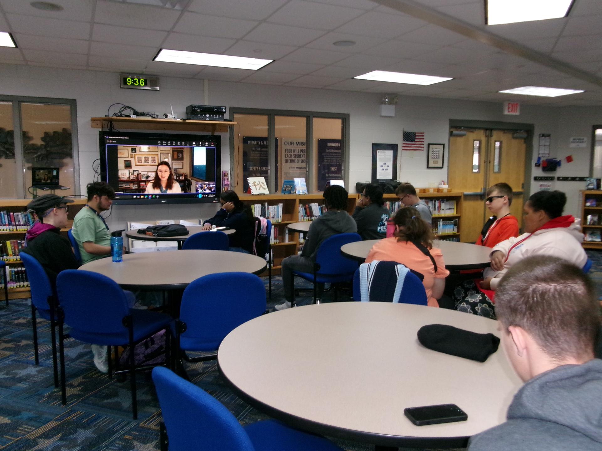 Students sit around the tables and chairs in the Blind Library; author Morgan Matson is on the screen