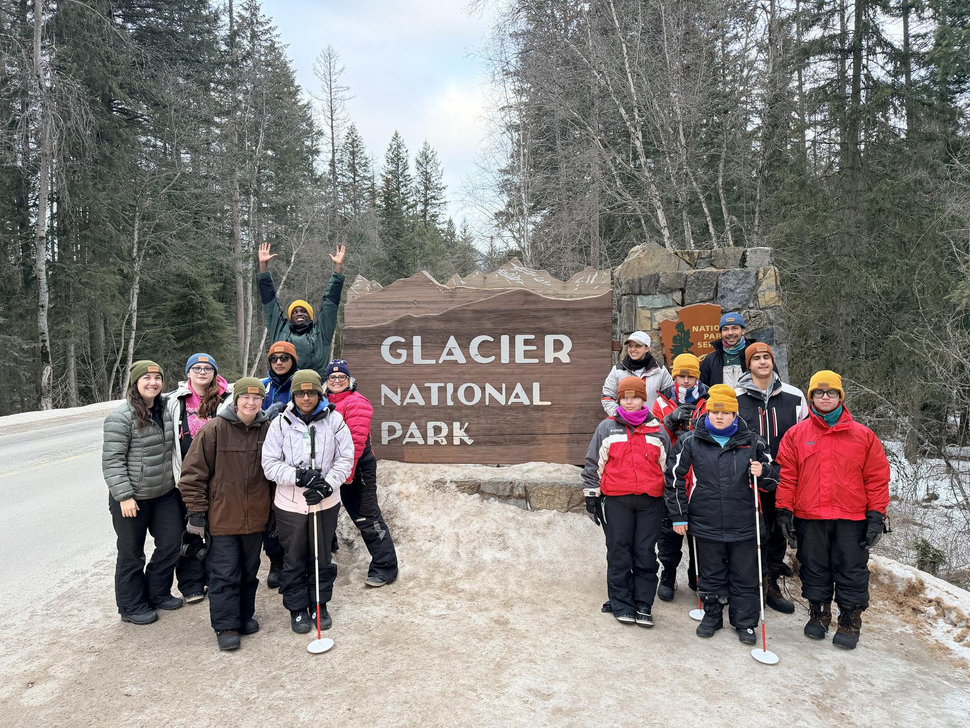 All students standing alongside road on a pile of old snow. They are standing in front of a Glacier National Park road sign made of wood and stone. Tall trees are along both sides of the road