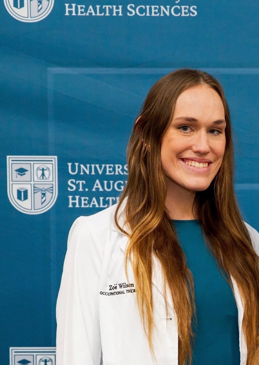 Zoe wearing her white coat standing in front of a University of St. Augustine Health Sciences backdrop