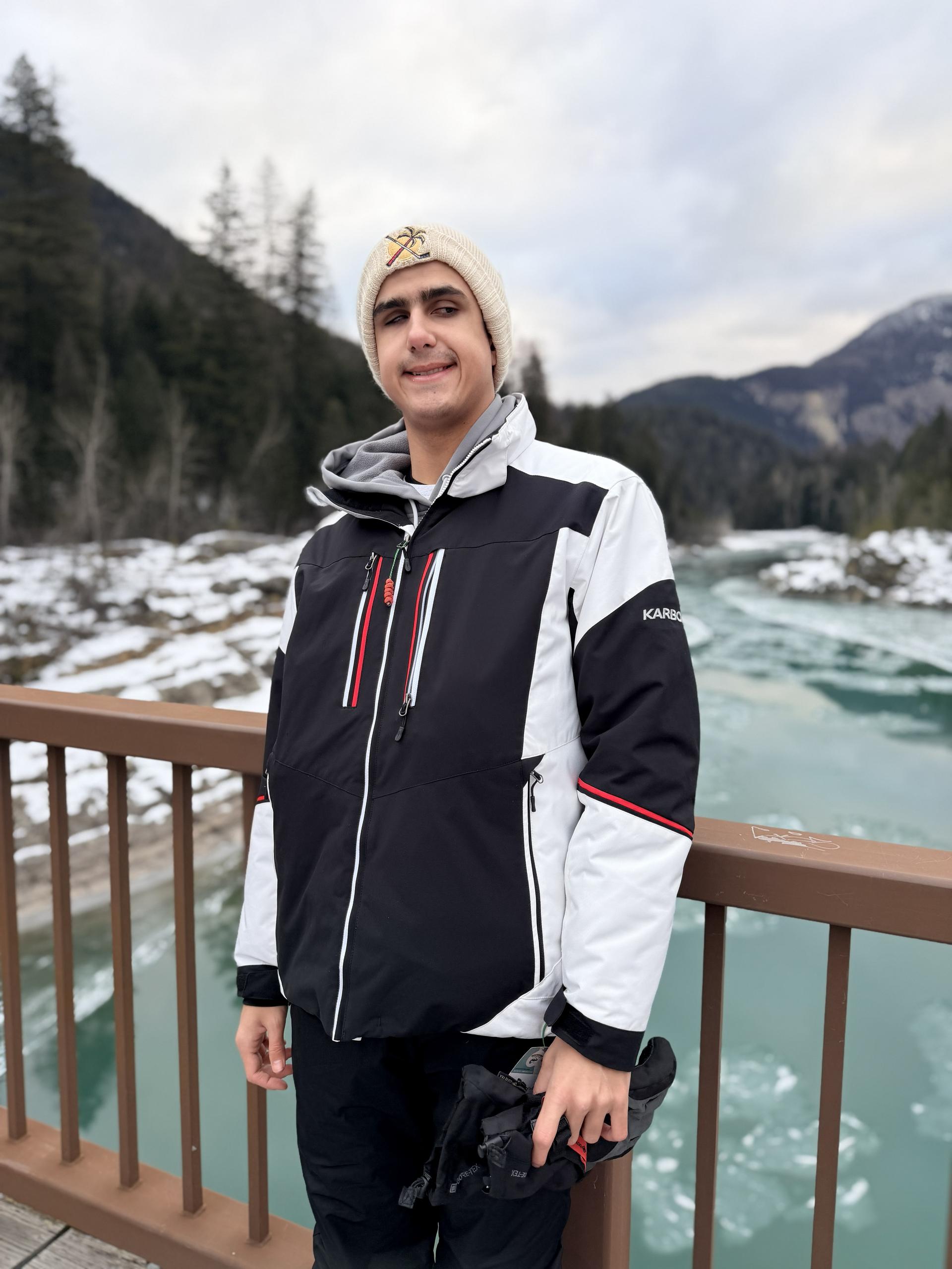 Damian. All the students are dressed in winter outdoor clothing. They stand on a bridge over a blue glacial river with ice and snow about. There is a mountain range in the distance, along with some tall spindly pine trees 