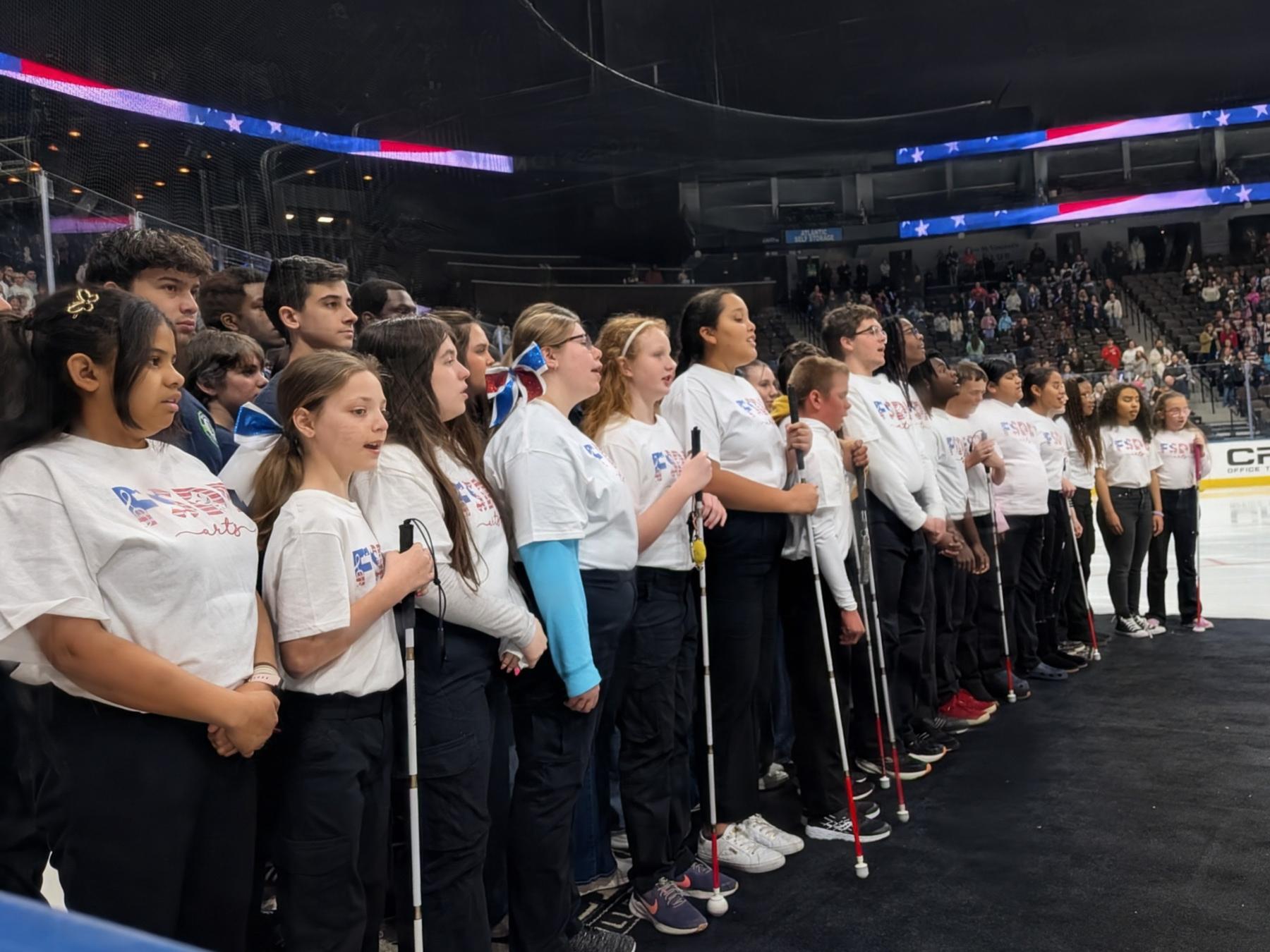 Show choir wearing black pants and white tshirts with a patriotic FSDB Arts logo standing in a line on the ice