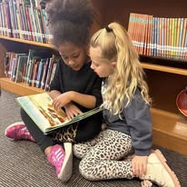 The image shows two young elementary school students sitting together on a carpeted floor in a library or classroom setting, looking at an open picture book together.