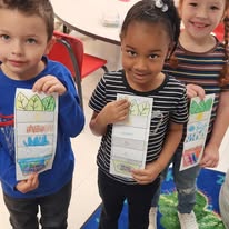 This photo shows three young students holding up paper plant diagram crafts (showing the parts of a plant, such as leaves, stem, roots, and pot/soil).  Due to the low resolution and distance, the handwritten student labels and printed text on the paper craft panels are too small and blurry to read clearly. However, the printable template sections typical for this activity include:  Leaves (top section)  Stem (middle section)  Roots / Soil / Pot (bottom section)