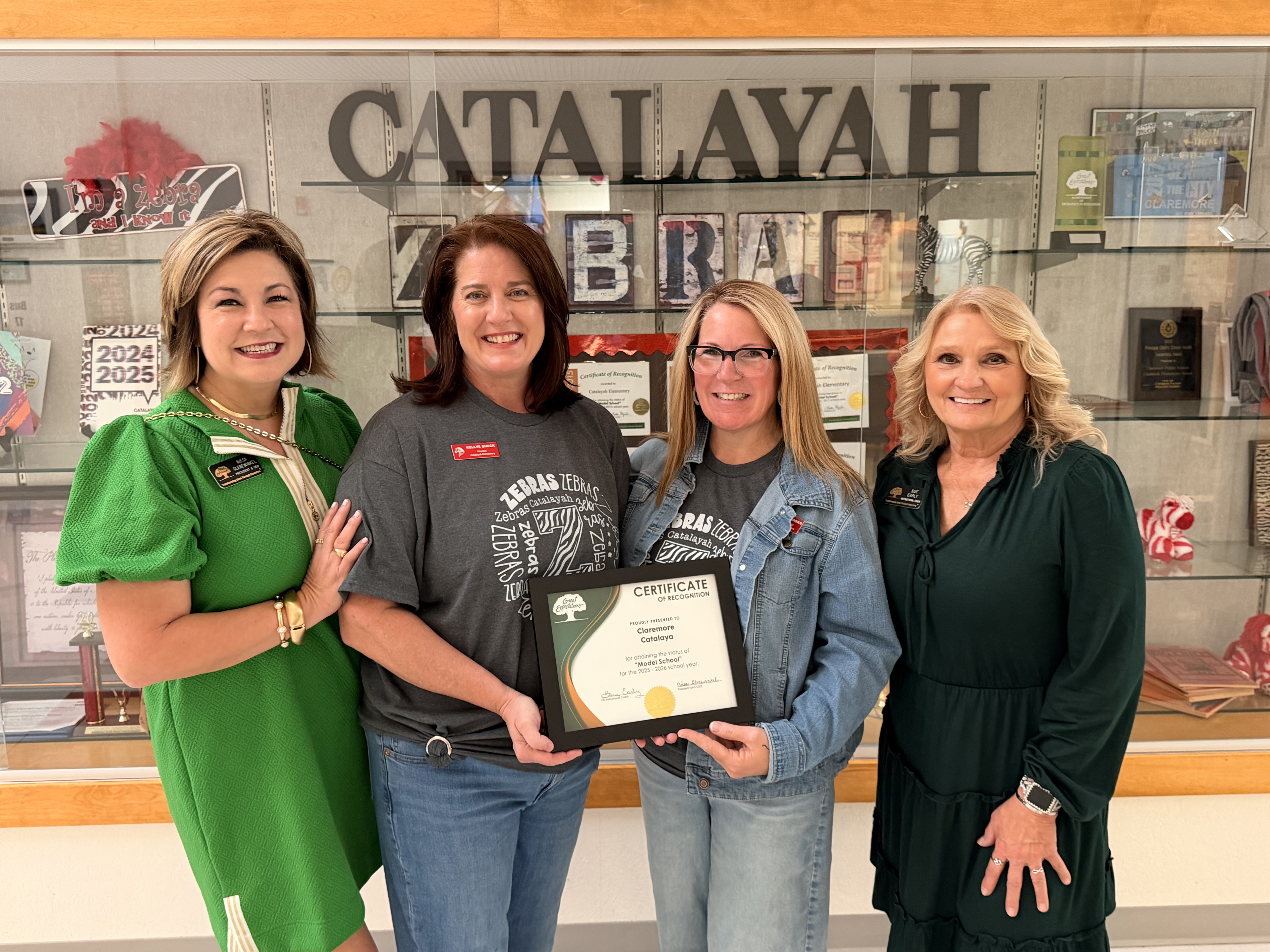 This photo captures a group of four women standing in front of a trophy case at Catalayah Elementary School, holding an award certificate. Certificate of Recognition The certificate held in the center of the photo reads: Organization: Great Expectations Awarded To: Claremore Catalayah Achievement: For attaining the status of 