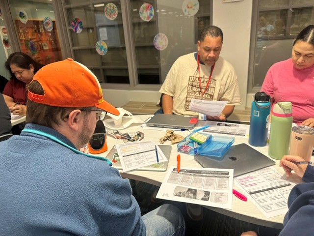 Group of people gathered around a table with laptops, papers, and pens. One person wears a red cap.