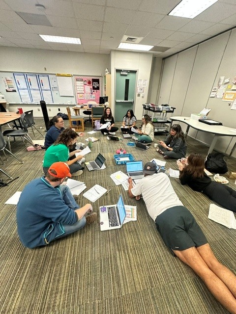Multiple individuals sit and lie on a carpeted floor in a room, engaged in a discussion with laptops.