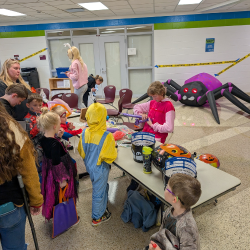 Children dressed in Halloween costumes gather around a table with games and treats, while a large inflatable spider decorates the background of the festive indoor event.