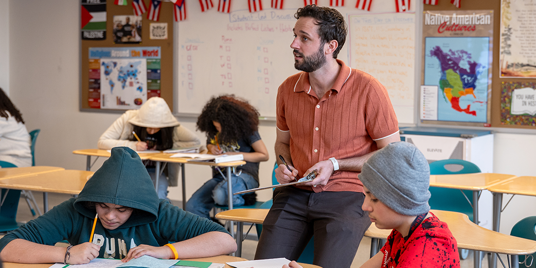 A teacher holds a clipboard and pen while working with students
